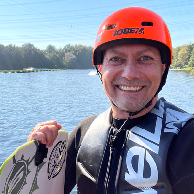 Spartenleiter Luke mit orangenem Helm und Prallschutzweste hält ein Wakeboard in der rechten Hand. Im Hintergrund ist ein See und Bäume zu sehen.